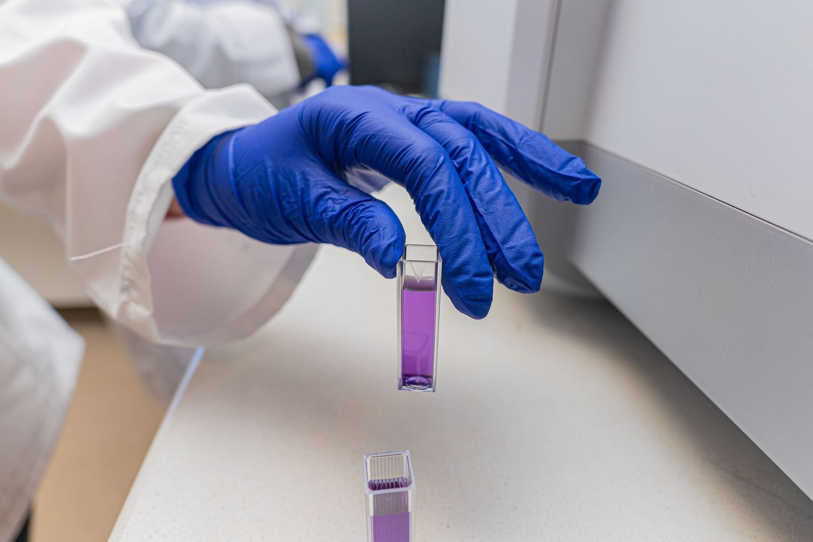 Gloved hand holding a cuvette of purple liquid at a laboratory bench