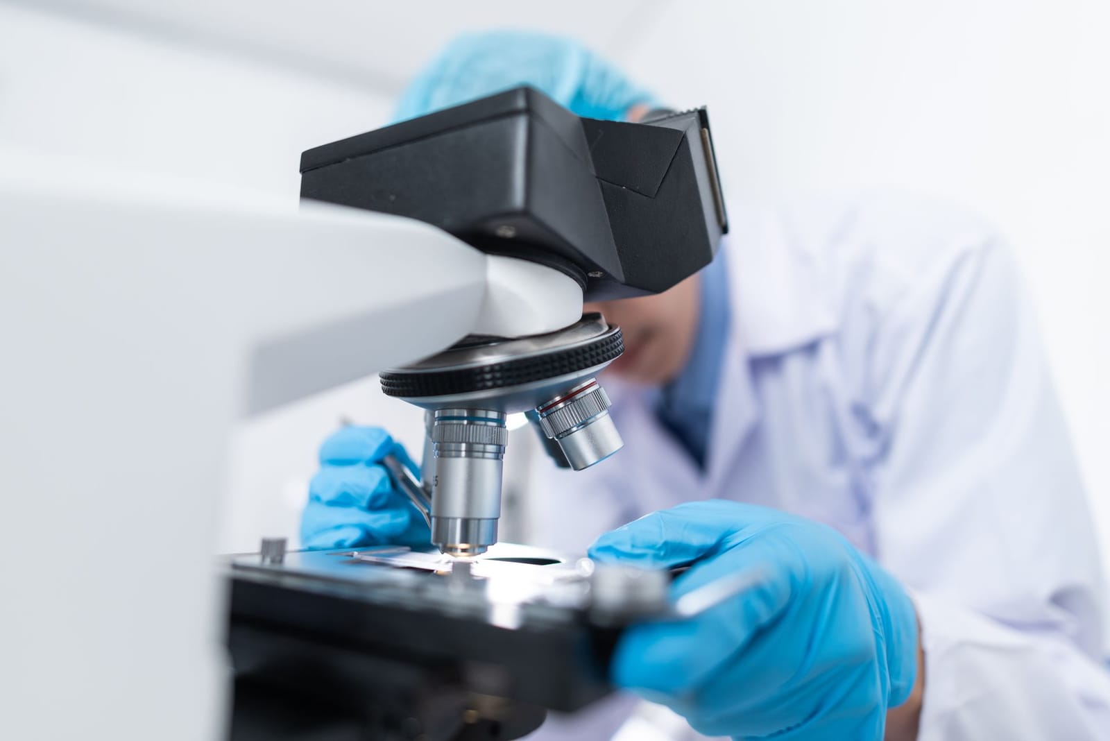 Scientist in a lab coat and blue gloves adjusting a microscope in a bright laboratory